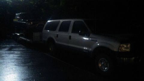1978 Ford Bronco - A white SUV is parked on a wet asphalt surface at night, with its headlights illuminated. Another dark vehicle is partially visible on a tr…