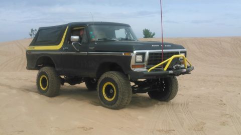 1978 Ford Bronco - A lifted, dark-colored Ford Bronco with yellow accents is parked on a sandy dune under a cloudy sky. The vehicle features a yellow brush gu…