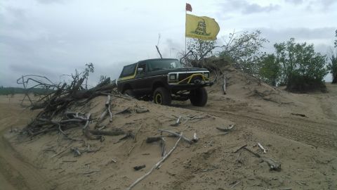 1978 Ford Bronco - A black 1978 Ford Bronco with a yellow stripe and aftermarket yellow front bumper is driving up a sandy dune. A yellow flag with a snake an…