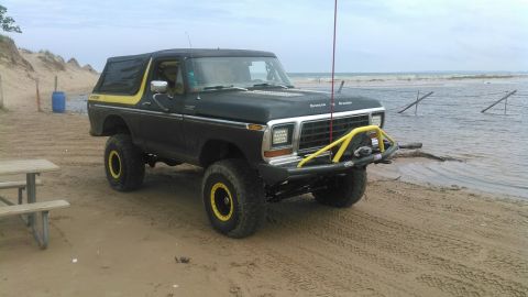 1978 Ford Bronco - A modified black 1978 Ford Bronco with yellow accents sits on a sandy beach near the water. The vehicle has large tires, a custom front bum…