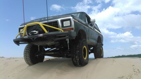 1978 Ford Bronco - A lifted, dark-colored 1978 Ford Bronco is parked on a sand dune with a blue sky and scattered clouds overhead. The vehicle features a yell…