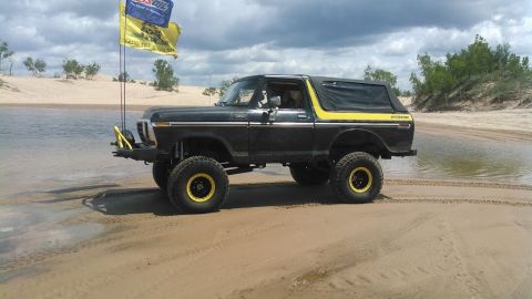 1978 Ford Bronco - A black 1978 Ford Bronco with yellow accents and large off-road tires is parked on a sandy shore next to a body of water. Two flags, one bl…