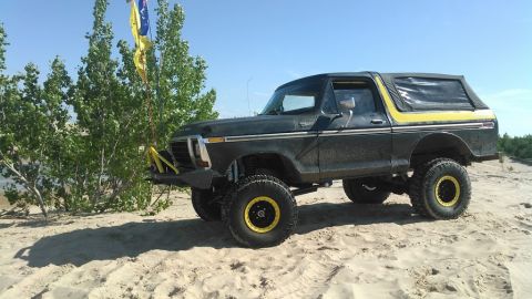 1978 Ford Bronco - A lifted, dark green 1978 Ford Bronco with yellow accents sits on a sandy dune under a clear blue sky. The vehicle has large off-road tires…