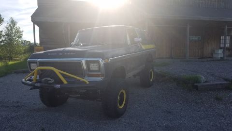 1978 Ford Bronco - A lifted, dark-colored Ford Bronco with a yellow brush guard and yellow accents on the wheels is parked in front of a wooden building with…