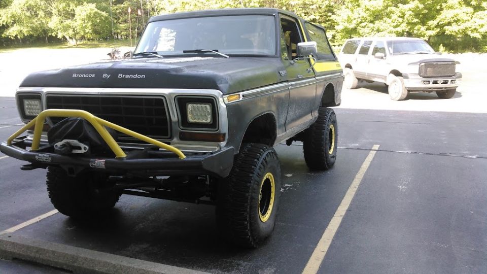 Lifted black 1978 Ford Bronco with yellow roll cage and custom bumper, parked next to a white SUV.