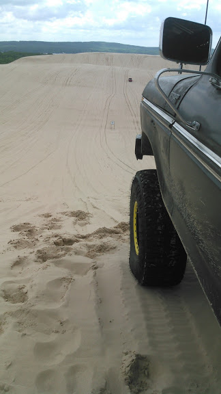 1978 Ford Bronco - A dark colored truck with a large off-road tire is parked on a sand dune overlooking a vast expanse of sand with tire tracks and a few dist…