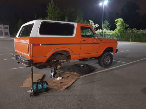 1978 Ford Bronco - This orange Ford Bronco is being worked on in a parking lot at night, with the rear wheel removed.