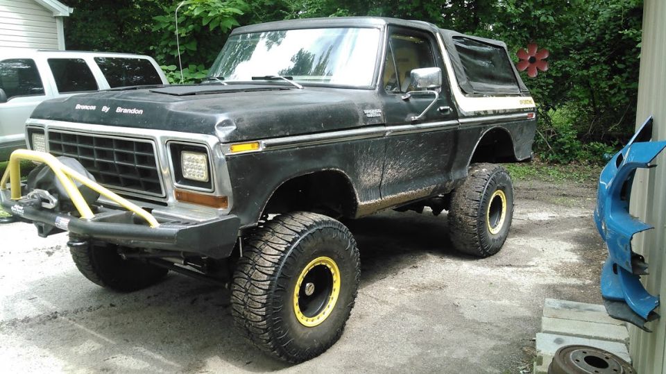 Black Ford Bronco with yellow accents on wheels and bumper, lifted with large tires. Another white SUV is parked behind it
