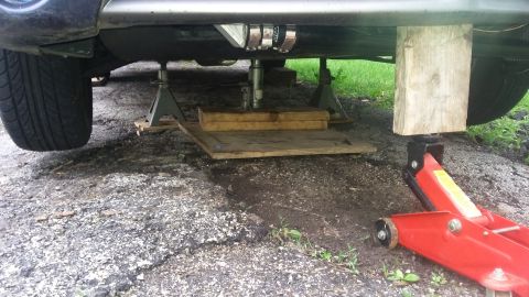 Car underside supported by jack stands and a red hydraulic jack, with wooden blocks. Tire visible on left