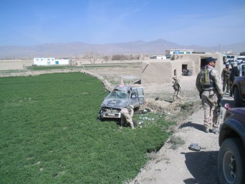 1981 Ford Bronco - A damaged sedan is stuck in a ditch next to a dirt road, with soldiers nearby and buildings in the background.