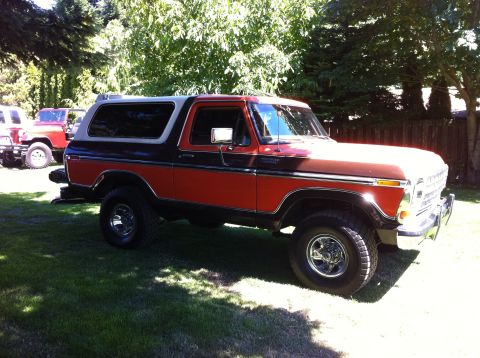 1978 Ford Bronco - This two-tone red and black Ford Bronco is parked on a grassy lawn with trees and a fence in the background.