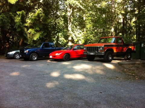 1978 Ford Bronco - Four vehicles are parked in a gravel lot with trees in the background.