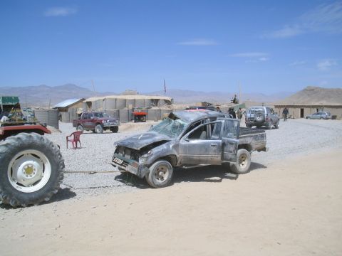 1981 Ford Bronco - A severely damaged pickup truck is being towed by a tractor in a dusty, arid environment with military-style fortifications.