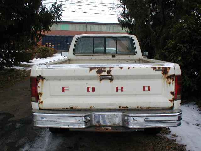 Rear view of a white Ford pickup truck with rust on the tailgate and red 'FORD' lettering