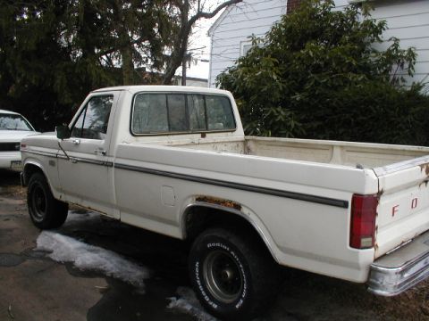 1986 Ford F-150 - A white pickup truck is parked next to another vehicle, showing its side profile with visible rust.