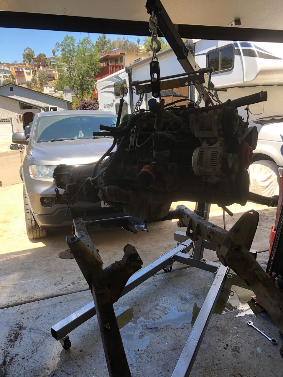 Engine and transmission assembly suspended by engine hoist, with a silver SUV in the background