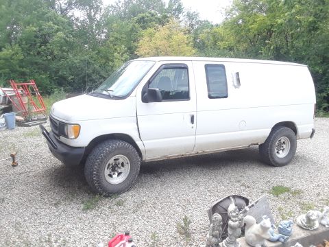1999 Ford E250 - A white 1999 Ford E250 van is parked on a gravel surface with trees and some construction materials visible.