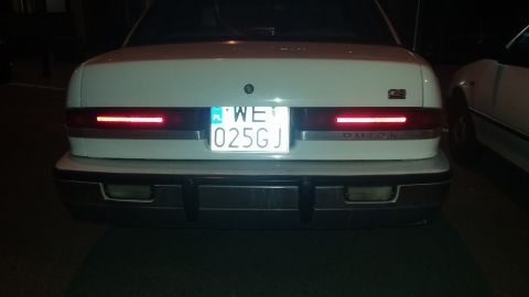Rear of a white car with a license plate reading WE 025GJ and 'BUICK' text on the trunk lid. Another car is pa