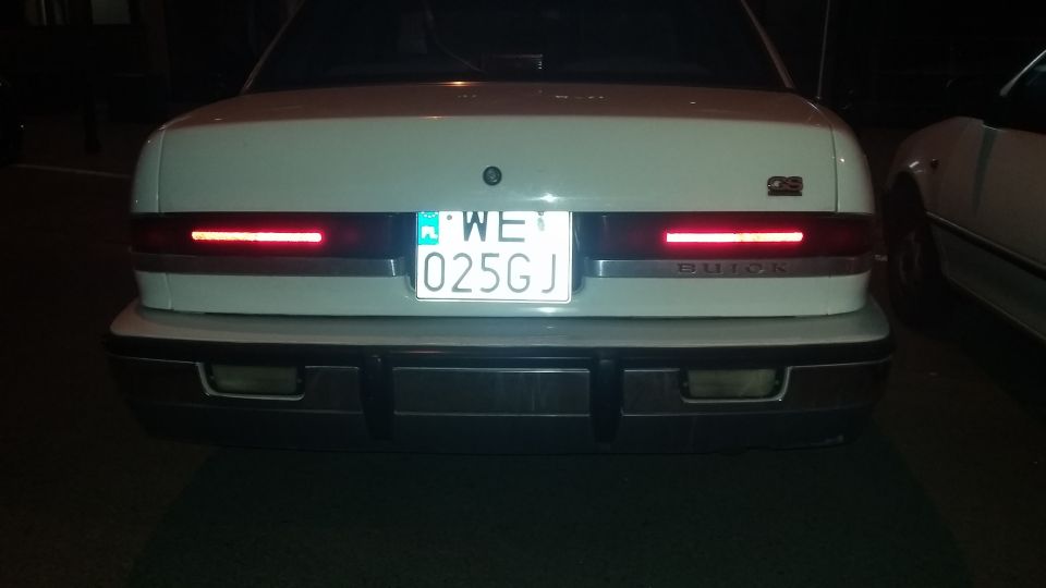 Rear of a white car with a license plate reading WE 025GJ and 'BUICK' text on the trunk lid. Another car is partially visible to the right