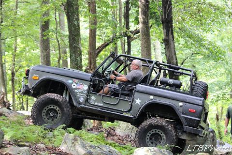 1972 Ford Bronco - A man drives a modified Bronco off-road through a dense forest with large rocks and foliage.