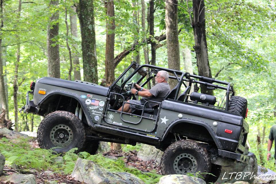 Dark green off-road Bronco with roll cage, driven by a man, navigating a rocky, wooded trail