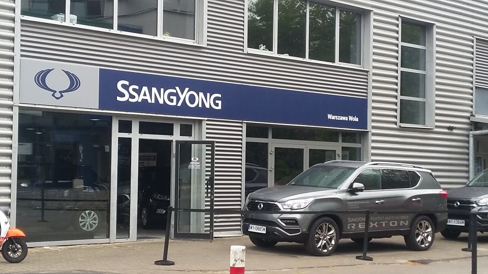 Gray SsangYong Rexton SUV parked in front of a dealership, showing "Rexton" text on the door