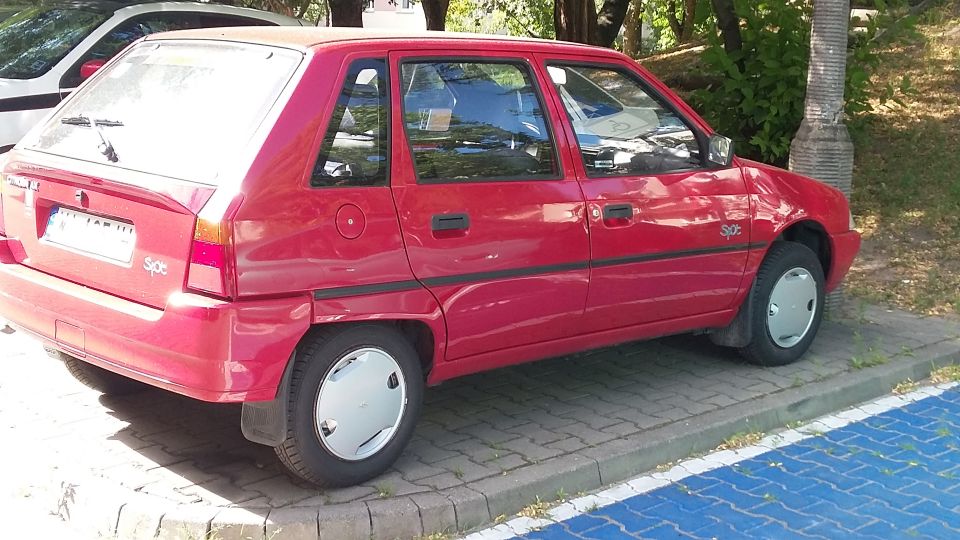 Red five-door hatchback car parked at a curb under trees, rear three-quarter view