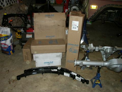 1995 Ford Bronco - Automotive parts, including leaf springs and a differential, are laid out with boxes and equipment in a garage.