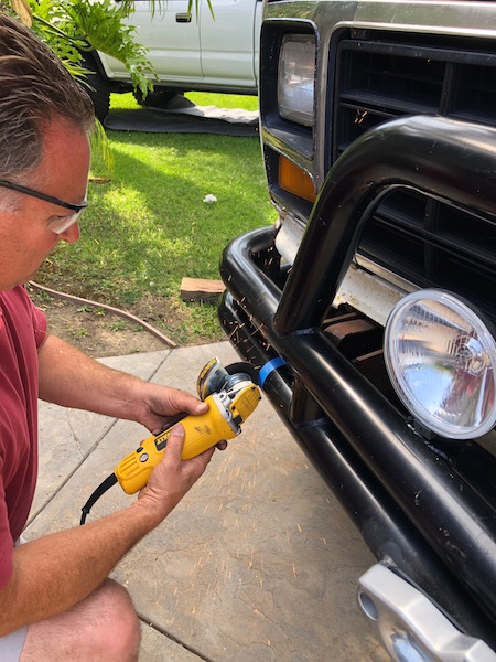 1981 Ford Bronco - A man uses an angle grinder on the bull bar of a truck, with another vehicle visible in the background.
