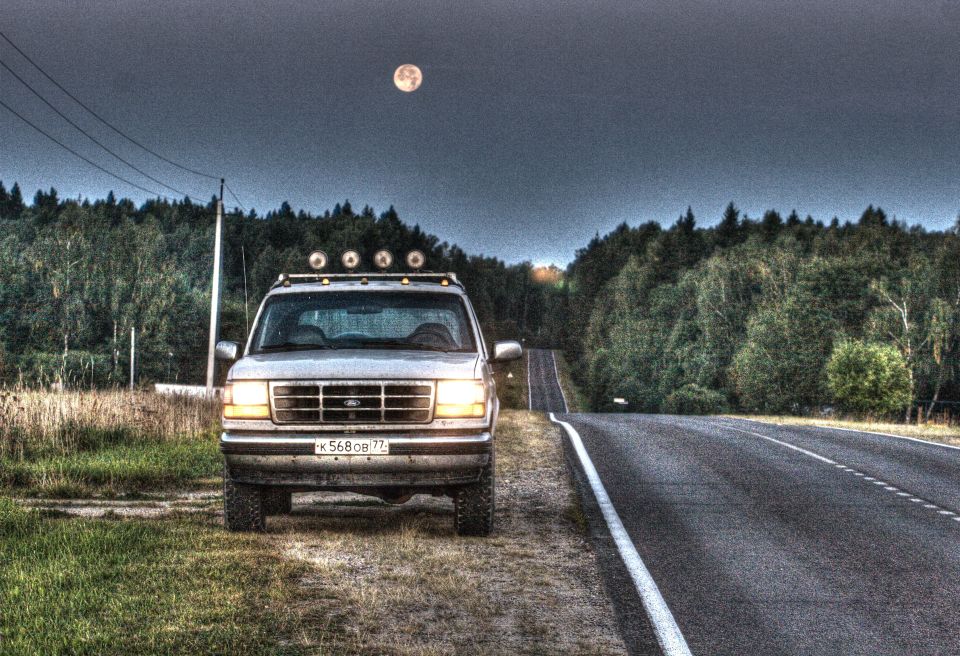 Front view of a 1993 Ford Bronco with a light bar on the roof, parked on a grassy shoulder next to a road