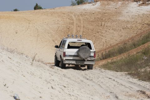 1993 Ford Bronco - A white Ford Bronco navigates a sandy terrain, kicking up dust as it ascends a hill.