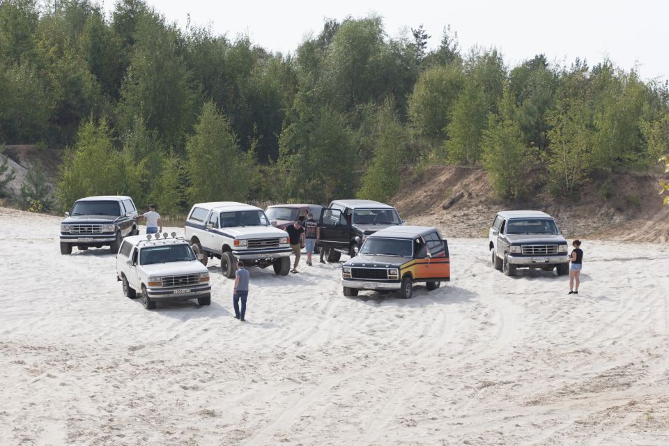 Several Ford Bronco SUVs parked on sand with trees in the background. One Bronco has an orange and yellow stripe
