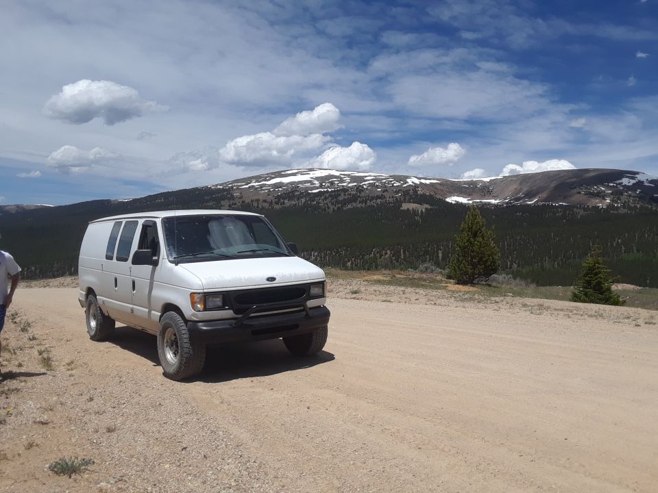 1999 Ford E250 van parked on a dirt road, mountains and cloudy sky behind it