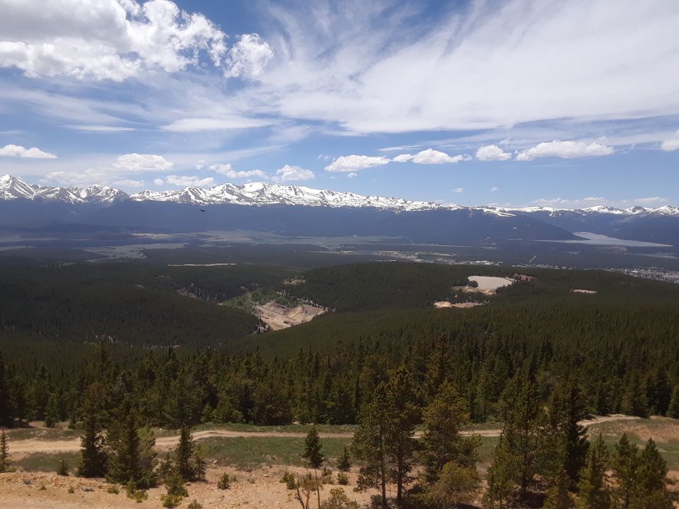 Mountain range with snow-capped peaks overlooks a vast forest and valley under a blue sky with clouds. A dirt road is visible in the foreground