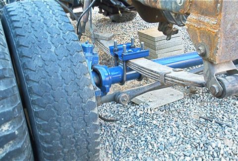 1986 Ford Bronco - Close-up of a freshly painted blue front axle and leaf spring suspension under a vehicle, viewed from the side near the tire. The setup res…
