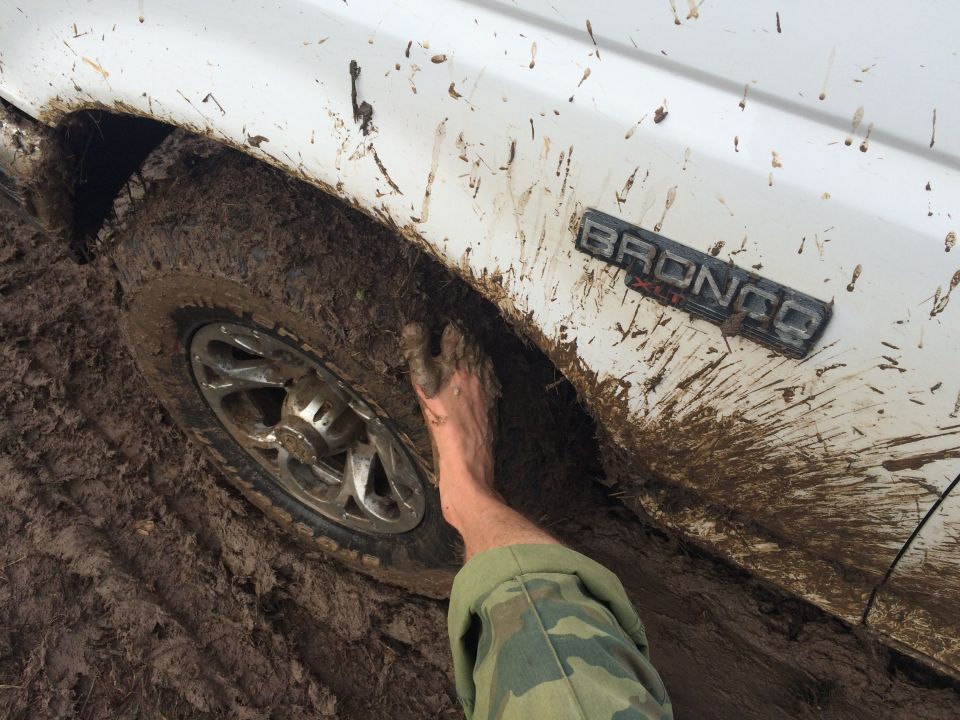 White 1993 Ford Bronco side view, covered in mud, with a foot stuck near the wheel