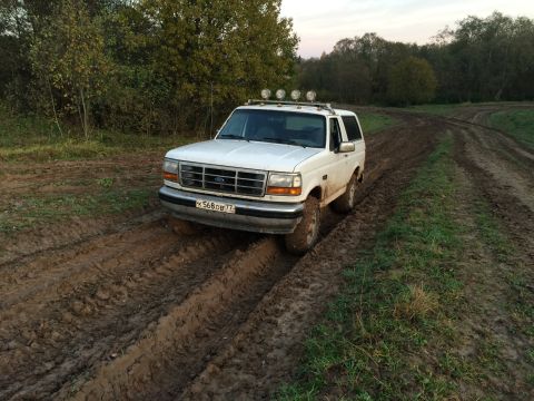 1993 Ford Bronco - White Ford Bronco navigates a muddy off-road path with trees in the background.