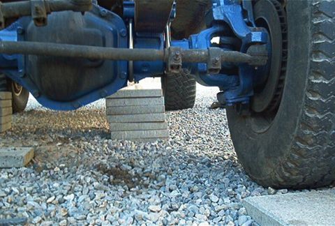 1986 Ford Bronco - Low-angle underside view of a front axle painted blue, showing the differential housing, tie rod, and steering knuckle, with a large off-ro…