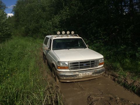 1993 Ford Bronco - A white 1993 Ford Bronco navigates a muddy off-road trail, its roof lights illuminated.