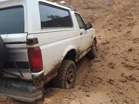 1993 Ford Bronco - A white 1993 Ford Bronco is stuck in a sandy terrain, viewed from the rear.