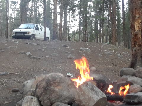 1999 Ford E250 - A white van sits in a wooded area near a campfire, suggesting a remote camping or off-road excursion.