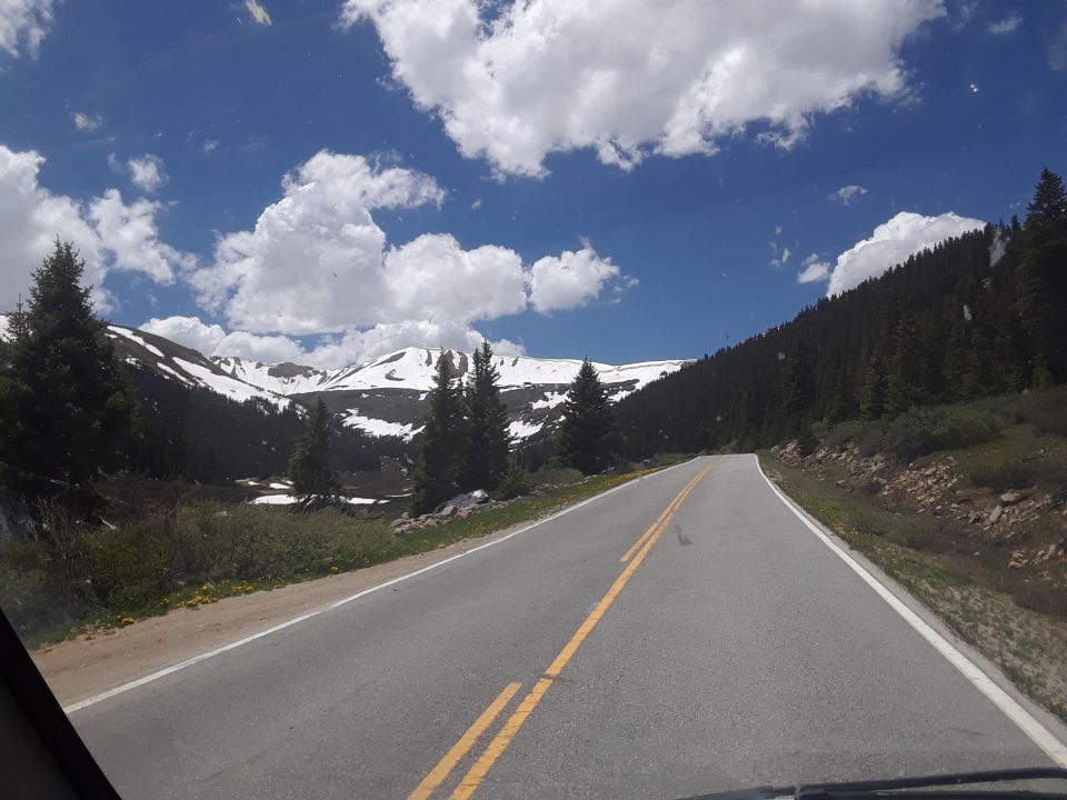 Mountain road with snow-capped peaks and pine trees under a blue sky with clouds. Supermotors.net logo in lower right