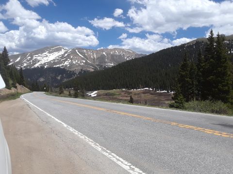1999 Ford E250 - A scenic mountain road winds through a forest, with snow on the distant peaks under a partly cloudy sky.