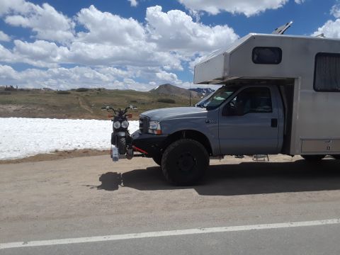 1999 Ford E250 - A truck camper is equipped with a front-mounted rack carrying a small motorcycle, seen on a scenic mountain pass.