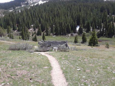 1999 Ford E250 - A rustic log cabin sits in a grassy clearing, surrounded by a dense pine forest and a distant mountain range.