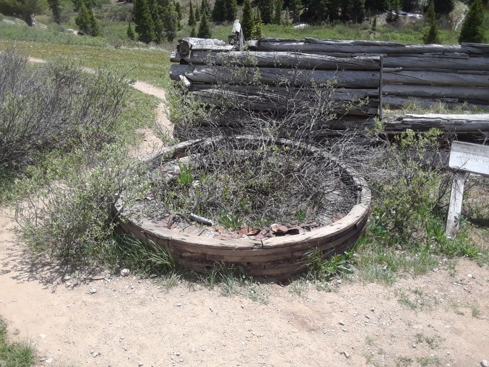 Wooden circular structure filled with brush, next to a log wall and a sign in a grassy, wooded area