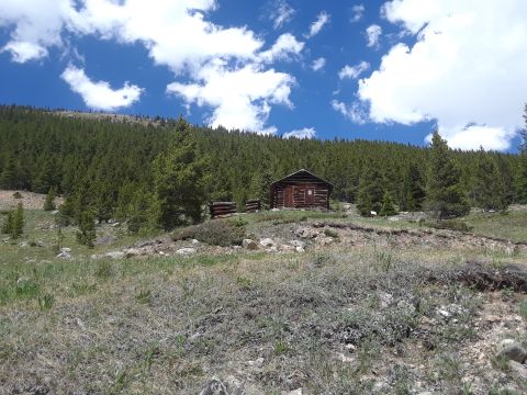 1999 Ford E250 - A rustic log cabin sits amongst pine trees on a mountainside, under a bright blue sky dotted with fluffy white clouds.