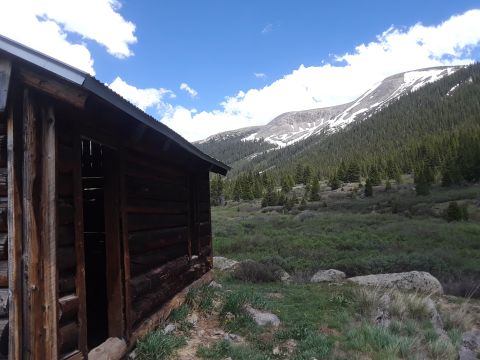 1999 Ford E250 - A rustic wooden cabin stands in a remote mountain valley, surrounded by dense pine forests and rocky terrain.