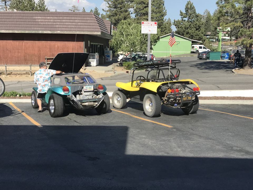 Teal dune buggy with open hood and a person nearby, parked next to a yellow dune buggy with a roof rack