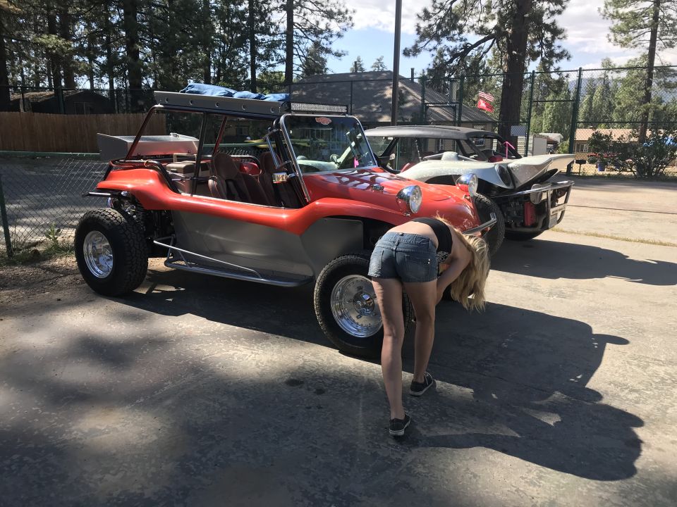 Red and silver dune buggy parked next to another vehicle, woman bending down to inspect the front wheel